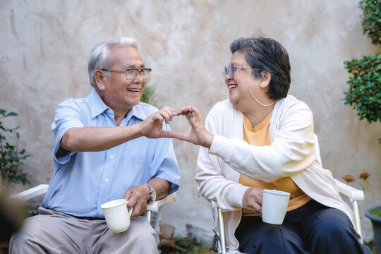 Elderly couple sits in garden chairs. Each holds a white teacup. They smile, joining hands in a heart shape. Their joy is clear and genuine. Ideal for retirement or family moments