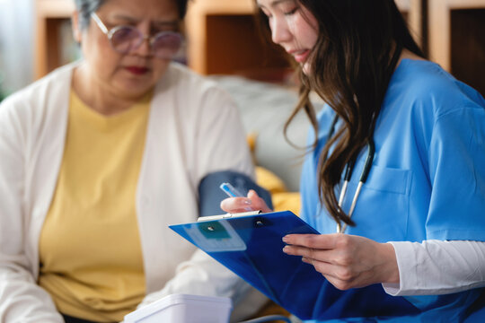 Healthcare professional in blue scrubs uses a blood pressure cuff and holds a clipboard with notes. Patient sits concern on couch. Nurse shows focus and care. The patient appears calm