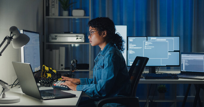 Young African American female programmer coding AI software on multiple screens in night office. Network security, cybersecurity, machine learning, working late, overtime, robotics automation concept.
