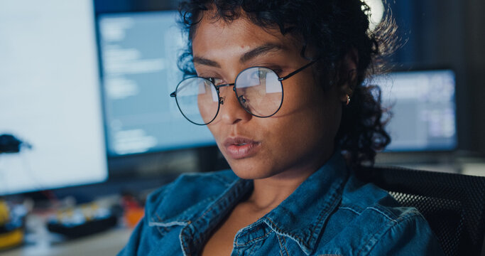 Close-up young Black female programmer coding AI software on multiple screens in night office. Network security, cybersecurity, machine learning, working late, overtime, robotics automation concept.