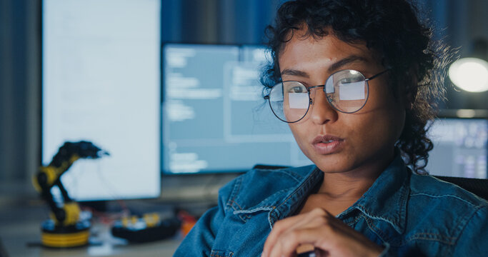 Close-up young Black female programmer coding AI software on multiple screens in night office. Network security, cybersecurity, machine learning, working late, overtime, robotics automation concept.