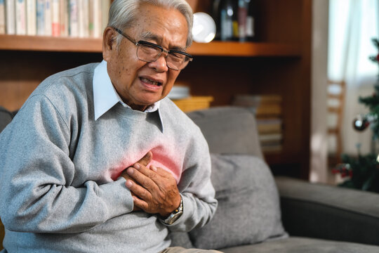 Elderly man holds his chest tightly. He shows clear pain and discomfort. His expression is distressed and worried. He sits on a gray sofa. Background includes bookshelves in living room at home