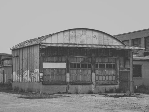 A black and white view of an old abandoned industrial warehouse loading dock building with multi pane windows, faded graffiti, and a curved metal roof