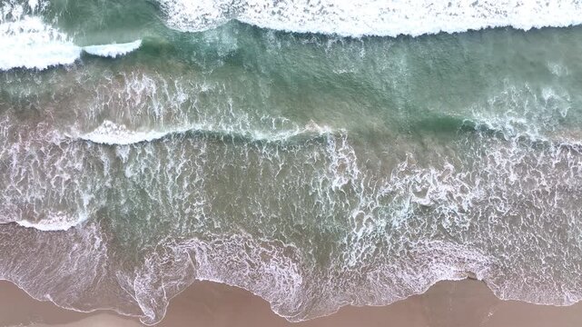 White foam spreading on beach from drone. Drone view of crashing foam waves. Sea foam patterns from aerial shot. Drone flying over foamy coastline. Ocean foam washing the sand from above.