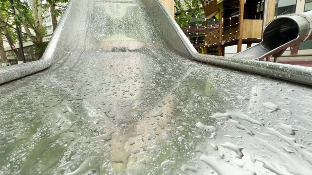 Rainwater droplets coat a shiny metal slide after a shower. The wet playground structure stands empty in a public park in Simferopol.