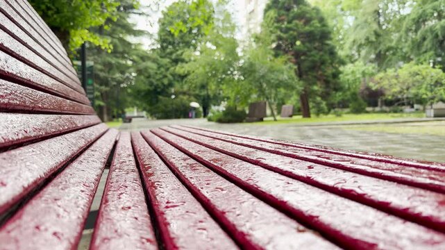 A wet red bench sits covered in raindrops. In the background, a person with an umbrella walks through the green public park. The ground and trees are damp from the rain.