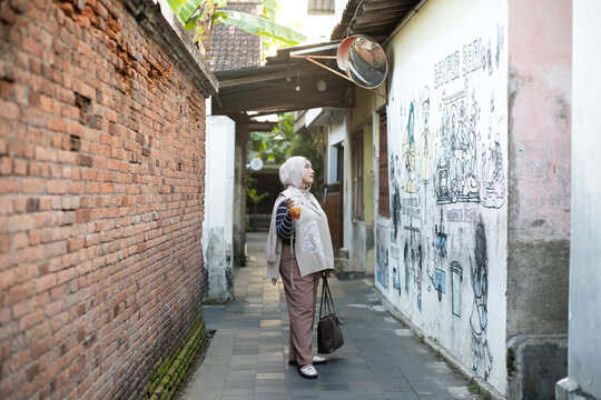 Full Length Portrait of Asian Muslim Woman Standing in a Traditional Javanese Alleyway Near Mural Wall