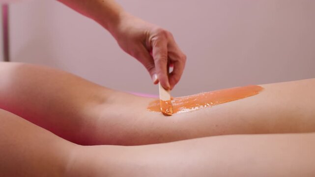 Hair removal process. Healthy treatment. Unrecognizable professional beautician hands waxing legs of woman with orange sugaring paste using spatula in spa salon.