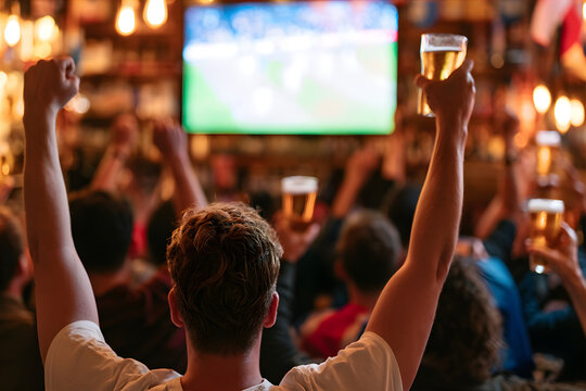 Man cheering while watching soccer championship in sport bar. Excited fan celebrating goal with raised arms and glass of beer. Group of people watching live game on television screen.