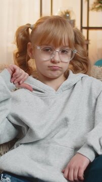 Young teenager girl showing thumbs down dislike with a disappointed expression at home. Caucasian child kid sitting on sofa, expressing dissatisfaction and disagreement through a clear hand gesture.