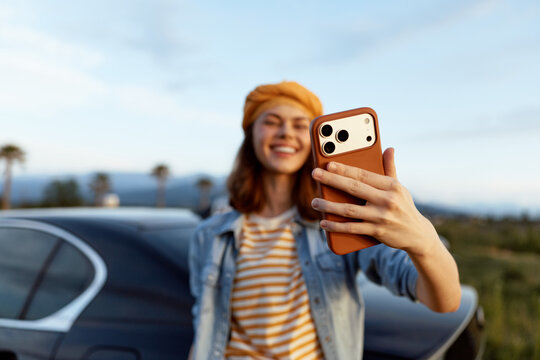 Young woman on a casual road trip takes a selfie with a smartphone, smiling beside a parked car, wearing a denim jacket and striped shirt, under clear blue sky for a carefree lifestyle moment