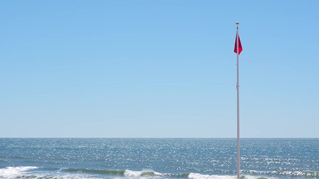 Red Flag Warning Waving Against Ocean. Signaling Hazardous Swimming Conditions. USA Panhandle No People. Wide Shot Water Sparkling Large Waves