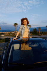 Fototapeta premium Young woman with curly hair stands through a sunroof, smiling, wearing a striped shirt and denim jacket in a bright outdoor scene with palm trees, clear sky and distant mountains.