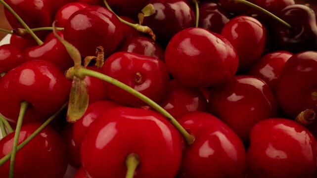 Rotating and panning closely over a pile of wet glossy fresh red sweet cherries glistening with water droplets as more cherries are falling onto the stack creating a textured background