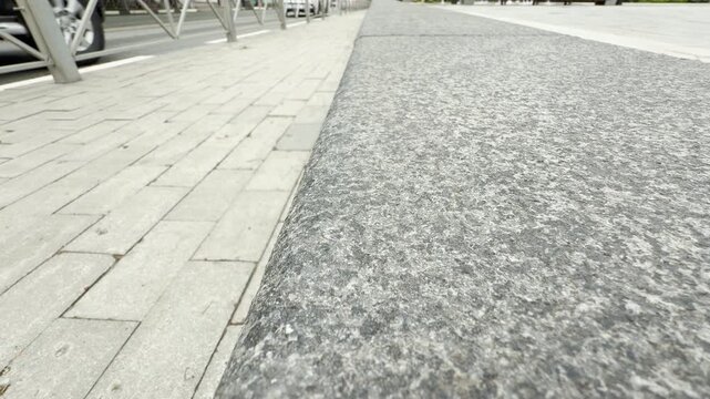 Cars scurry along the urban road behind a large stone curb. The paved sidewalk leads the eye toward the busy street in this city setting.
