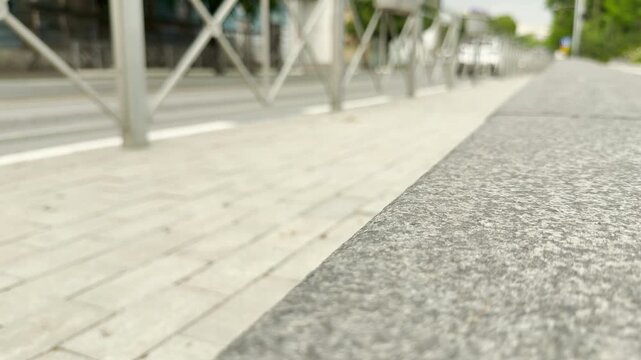 Cars scurry along the city road behind a metal fence and stone curb. The paved sidewalk leads the eye toward the blurred traffic in this urban setting.