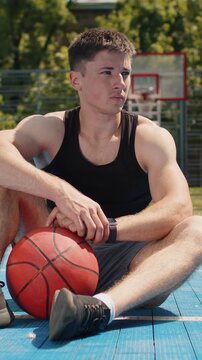 Portrait of tired athletic caucasian man in sportswear after playing basketball game. Boy sitting relaxing on urban city court, listening music, holding ball. Fitness routine on sports field. Outdoors