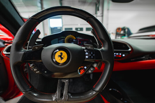 Close up interior view shows Ferrari 296 GTB carbon fiber steering wheel with red stitching, prancing horse emblem, paddle shifters, illuminated controls, and yellow rev counter in workshop.