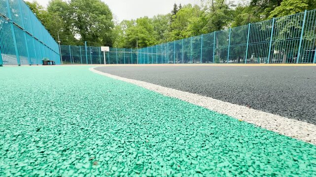 An empty basketball court sits in a peaceful public park. The textured green and grey surface waits for players to arrive and enjoy a game outdoors.