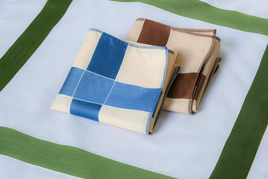A blue-and-beige and a brown-and-beige folded handkerchiefs neatly arranged together on top of a green-bordered gray cotton handkerchief.