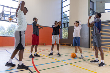 Fototapeta premium Diverse male teammates stretching arms across chest on school gym court near basketball, in shorts