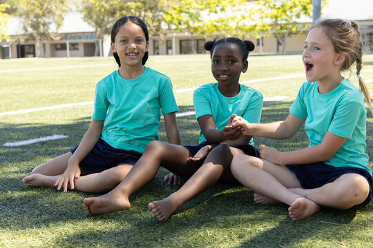 Diverse children sitting on school turf white line, wearing teal tees, navy shorts, bare feet