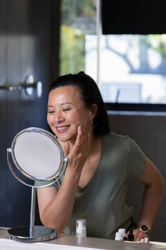 Asian woman sitting at bathroom vanity in olive shirt touching face checking mirror and bottles