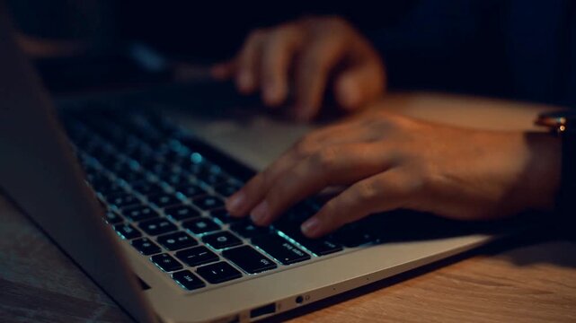 Cinematic close up of hands typing on laptop keyboard at night, person working late in dark room, programmer coding on computer with warm lighting