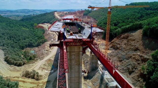 Aerial view of a viaduct construction site with concrete piers, deck formwork, crane operation and workers building an elevated road structure across a rural valley.