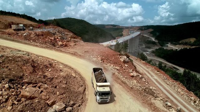 Dump trucks and drilling machinery working at a mountain excavation site, transporting soil and rock material through a dusty construction road during infrastructure development.