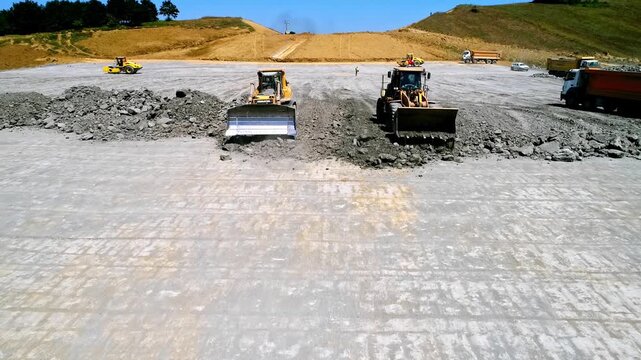 Heavy machinery spreading and leveling rock material at a large road construction site, with loaders, bulldozers, dump trucks and compactors preparing the road base.