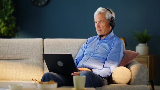 Elderly man using a laptop and headset during an online meeting or video conference from home. Remote communication, technology and active retirement.