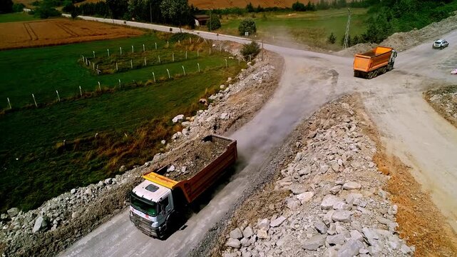 Aerial view of dump trucks carrying gravel and soil on a rural road construction site, showing material transport, rocky terrain and infrastructure development in the countryside.