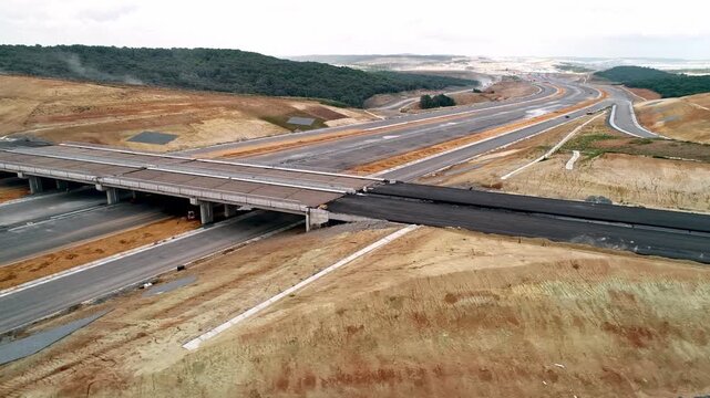 Aerial view of a highway construction project with bridge, asphalt roads and wide transportation infrastructure across rural landscape and forest area.