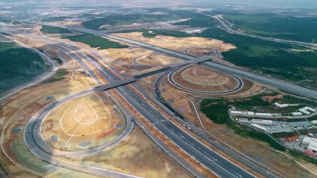 Aerial drone view of a large highway interchange with curved ramps, overpasses, traffic and surrounding forest, showing modern transportation infrastructure and road development.