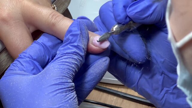 Nail technician works with precision and care. Hands are steady, focused, and skilled. Blue gloves protect and enhance cleanliness. Electric tool shapes nail with gentle control