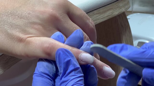 Nail technician files a client's nail with care. Blue gloves cover hands, showing professionalism. Ring gleams on client's finger, adding detail. Focused, gentle touch signals skill and comfort