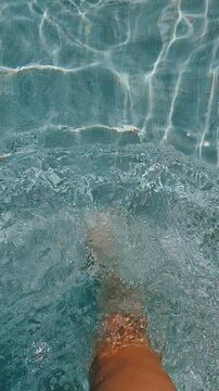 Bare legs splashing in clear blue swimming pool water over tiled bottom, relaxing summer vacation moment with sunlight reflections and gentle ripples