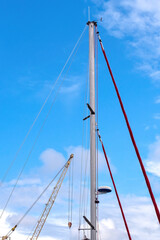 Fototapeta premium High sailboat mast with rigging and wires standing against a blue sky with clouds next to an industrial crane.