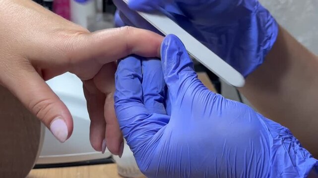 Nail technician files a client's nail with care. Blue gloves cover hands, showing professionalism. Ring gleams on client's finger, adding detail. Focused, gentle touch signals skill and comfort