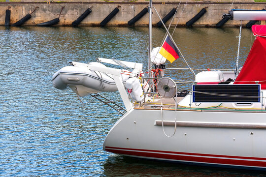 Stern of a cruising sailboat with a German flag, solar panel and an inflatable dinghy on davits in a port harbor.