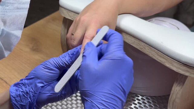 Nail technician files a client's nail with care. Blue gloves cover hands, showing professionalism. Ring gleams on client's finger, adding detail. Focused, gentle touch signals skill and comfort