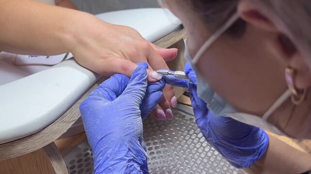 Nail technician works with precision and care. Hands are steady, focused, and skilled. Blue gloves protect and enhance cleanliness. Electric tool shapes nail with gentle control