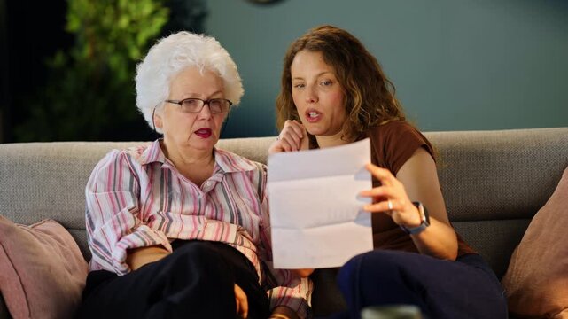 Adult daughter and elderly mother reviewing a household utility bill together at home. Family support, budgeting, financial planning and everyday expense management.