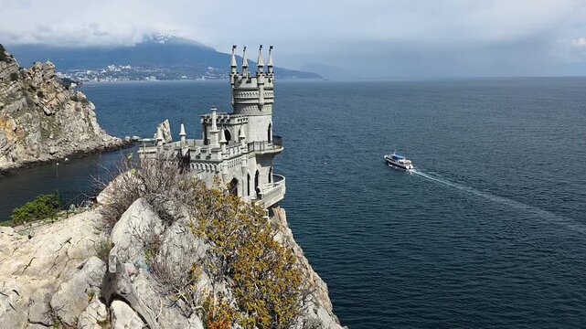 Swallow's nest castle, a neo-gothic landmark, standing perched on a dramatic rock cliff above the dark black sea, with a tour boat navigating the water creating a wake