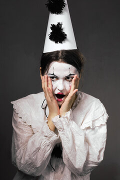 Shocked woman in clown makeup holding face in surprise wearing cone hat