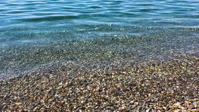 Shallow, transparent sea water with gentle ripples covering a colorful pebble beach, creating a beautiful underwater texture and tranquil seaside surface