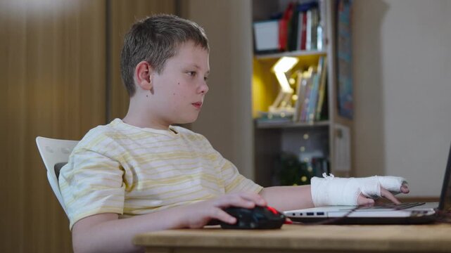 Caucasian boy with cast on arm plays on laptop and expresses emotions of excitement, surprise, and concentration. Teen gamer enthusiastically plays computer games despite injury.