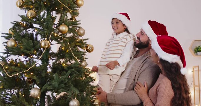 Christmas family in Santa hats decorating tree at home together. Father lifting daughter up to help hang golden ornaments on pine branch, parents and child decorate festive room with balls and garland