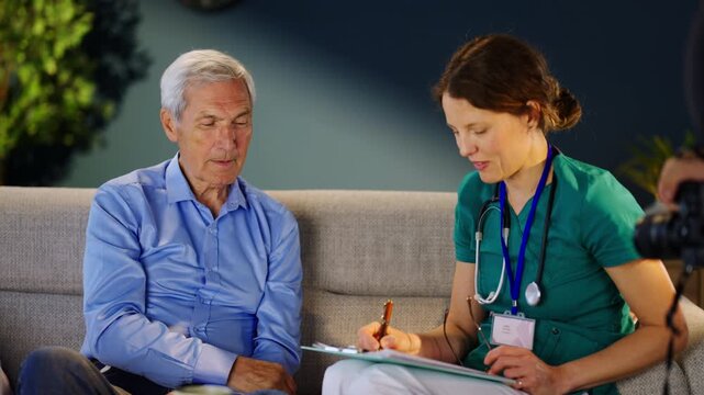 A doctor sits with an older patient in a clinic. The doctor takes notes while listening to the patient. They are discussing health concerns in a friendly way.